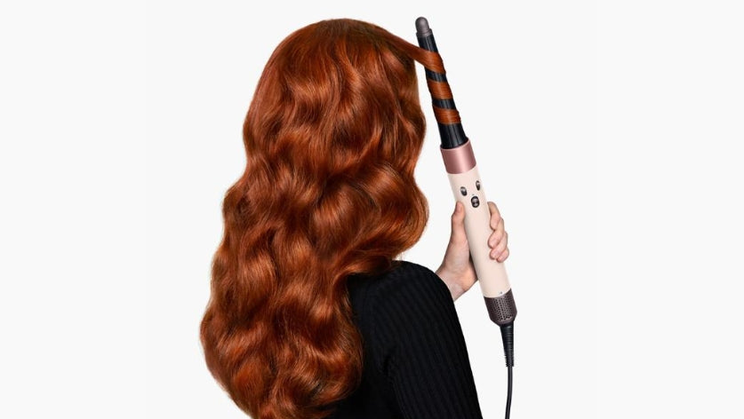 A woman with long, wavy red hair is shown from the back, using a curling iron to style her hair against a plain white background.