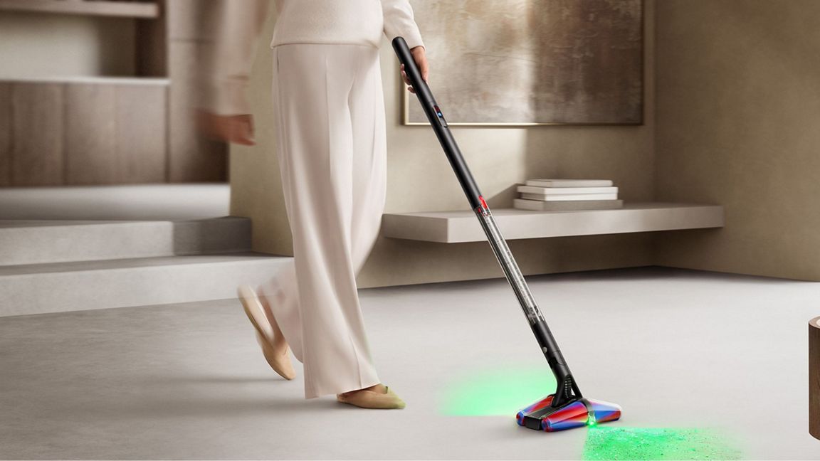 A person in a cream-colored outfit vacuums a light-colored floor in a modern interior setting, using a cordless stick vacuum with a colorful floor head that illuminates the floor with a green light.
