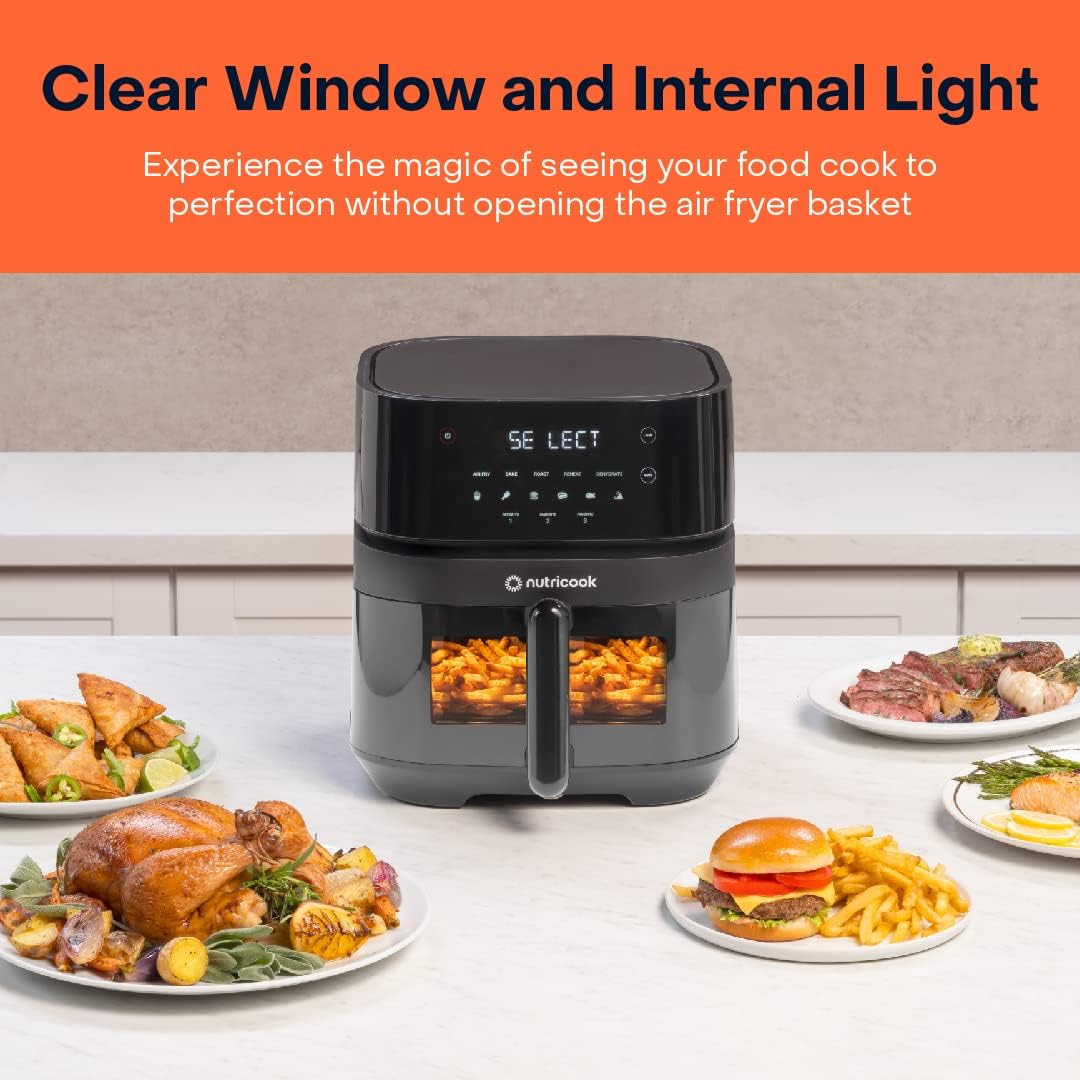 Black air fryer with clear window displaying food, surrounded by plates of cooked meals on a kitchen counter.