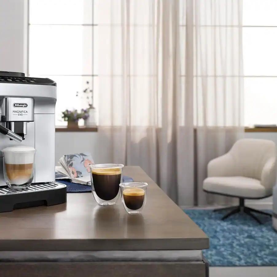 Taghareed De'Longhi Magnifica Evo automatic coffee machine in silver and black, brewing a latte. Two espresso cups are on the counter.