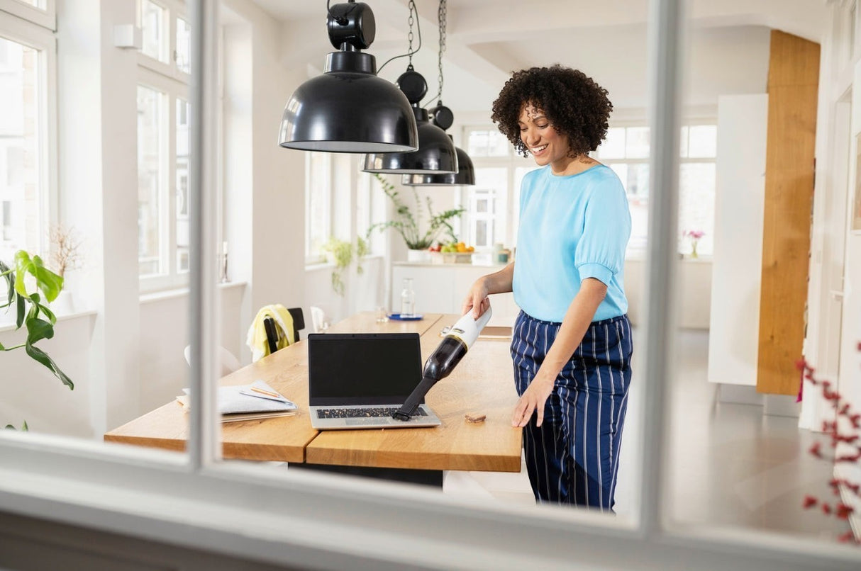 Taghareed Kärcher CVH 2 cordless handheld vacuum cleaner with HEPA filter, white and black, cleaning a laptop.