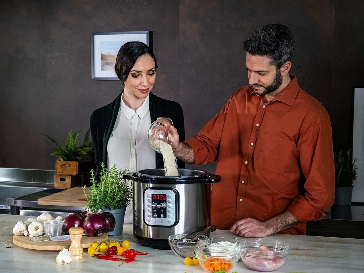 Taghareed Kenwood 16-in-1 8L electric pressure cooker in silver and black, with digital display, being used by a couple in a kitchen.