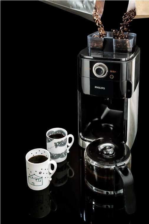 Taghareed Philips Grind & Brew coffee maker with built-in grinder, dual bean container, and glass carafe. Two mugs of coffee are beside it.