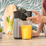 A Braun countertop juicer in black and stainless steel, with a hand pressing the dispensing button while orange juice fills a clear glass. The scene is set on a wooden kitchen counter with a patterned blue-gray backsplash in the background.