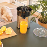 Braun juice press with a glass of orange juice on a table, surrounded by lemons and a dog in the background.