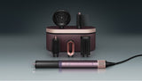 A hair styling tool set with a metallic purple and rose gold cylindrical wand, several black brush attachments, and a matching charging case on a dark gray gradient background.