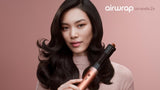 A woman with long, wavy dark hair holds a rose gold and black Airwrap Coanda 2x curling iron against a solid dusty pink background. The brand name 'airwrap' is visible in the top right corner.