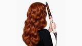A woman with long, wavy red hair is shown from the back, using a curling iron to style her hair against a plain white background.
