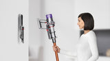 A woman in a white long-sleeve top holds a cordless stick vacuum cleaner with a metallic silver body, purple cyclone component, and copper-colored wand. The background is a clean, minimalist white wall with a small, out-of-focus reflection of the vacuum on the left.
