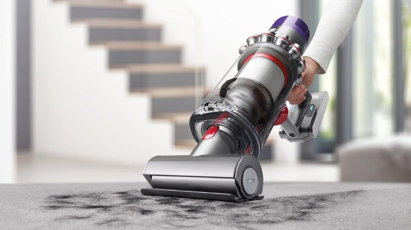 A cordless stick vacuum cleaner being held by a person on a gray carpet in a modern home interior, with stairs blurred in the background.