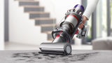 A cordless stick vacuum cleaner being held by a person on a gray carpet in a modern home interior, with stairs blurred in the background.
