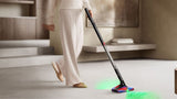 A person in a cream-colored outfit vacuums a light-colored floor in a modern interior setting, using a cordless stick vacuum with a colorful floor head that illuminates the floor with a green light.