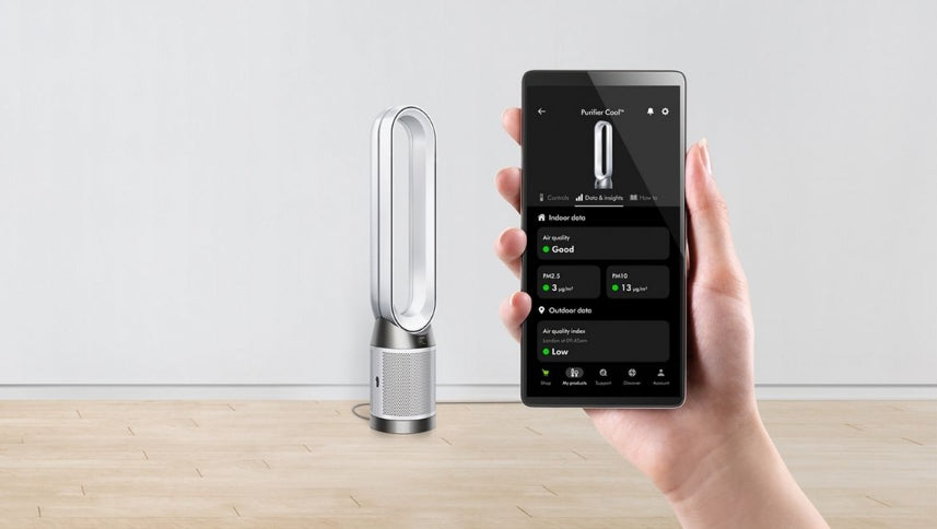 Hand holding a smartphone with app controls displayed next to a Dyson air multiplier on a wooden surface.
