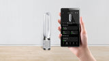 Hand holding a smartphone with app controls displayed next to a Dyson air multiplier on a wooden surface.