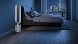 A tall, slim white and silver floor fan stands beside a bed in a modern blue-toned bedroom, with a person sleeping under white bedding and a city skyline visible through the window.