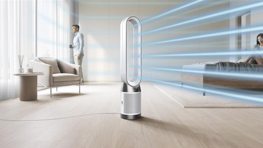 A sleek, modern floor-standing tower fan with a curved white and silver blade assembly stands on a light wood floor in a bright, contemporary living room. Blue graphic lines radiate from the fan to suggest airflow, with a man standing near a chair in the background and a woman sitting on a bed in the distance.