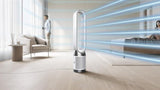 A sleek, modern floor-standing tower fan with a curved white and silver blade assembly stands on a light wood floor in a bright, contemporary living room. Blue graphic lines radiate from the fan to suggest airflow, with a man standing near a chair in the background and a woman sitting on a bed in the distance.