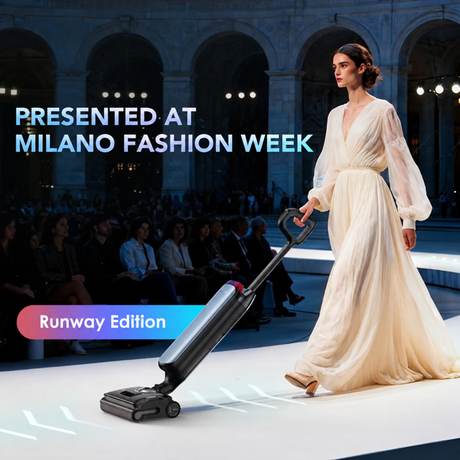 A woman in a flowing cream gown walks a fashion runway while pushing a sleek black and gray upright vacuum cleaner. Large text reads 'PRESENTED AT MILANO FASHION WEEK' and 'Runway Edition' over a dark arena with seated audience members in the background.
