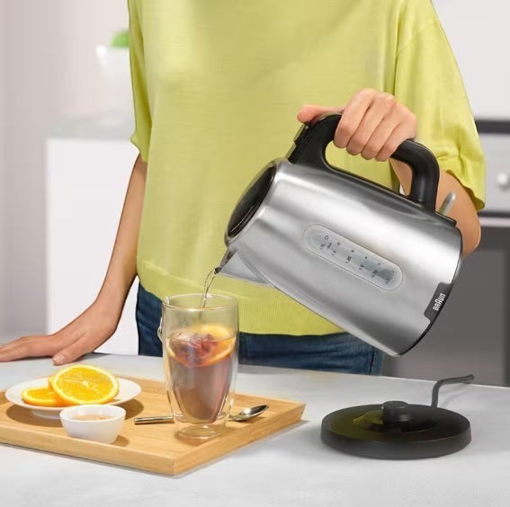 Braun PurShine stainless steel electric kettle pouring hot water into a glass with fruit slices. 1.7L capacity, 2200W.