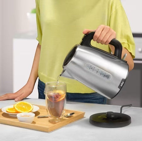 Braun PurShine stainless steel electric kettle pouring hot water into a glass with fruit slices. 1.7L capacity, 2200W.