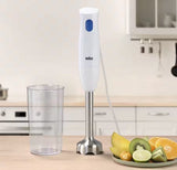 Taghareed Braun MultiQuick 1 hand blender, white with blue button, stainless steel shaft, next to a glass and fruit.