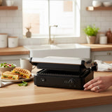 Taghareed CONTI Grill 2000W GT2201-G, black, with removable non-stick plates and 180° opening, shown in a kitchen setting with grilled sandwiches.