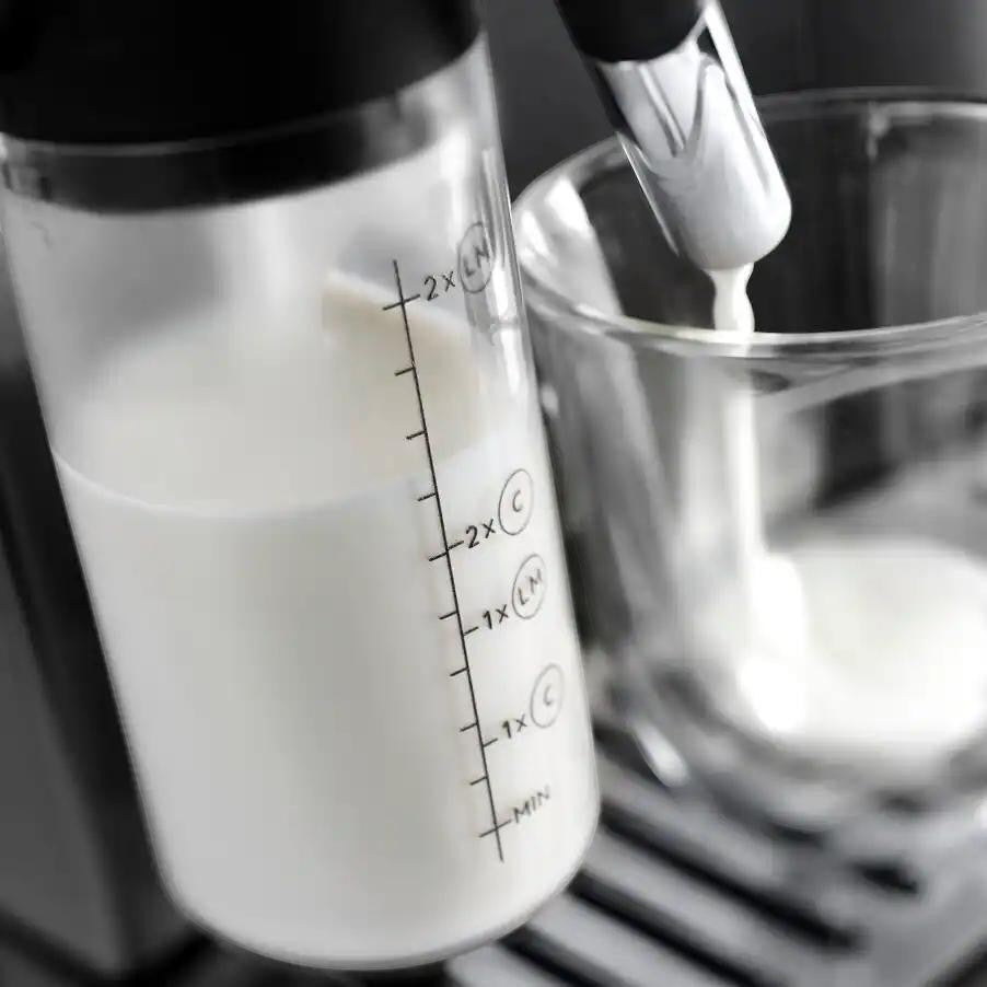 Taghareed De'Longhi Magnifica Evo automatic coffee machine frothing milk into a glass. Features a clear milk carafe with measurement markings.