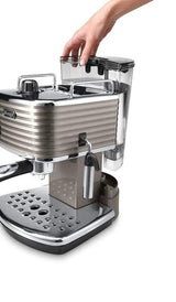 Taghareed De'Longhi Scultura espresso machine in metallic grey with a hand removing the water tank. Features a chrome drip tray.