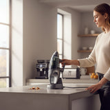 Taghareed Kenwood 14.8V cordless handheld vacuum cleaner, grey, with charging base and crumbs on counter.