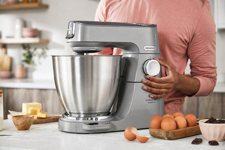 Taghareed Kenwood Titanium Chef Baker XL stand mixer in silver with a 7L stainless steel bowl and a person's hand adjusting the speed dial.